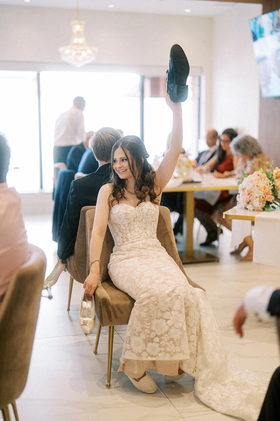 A bride in a lace wedding dress participating in a shoe game at a wedding reception, smiling and holding up a shoe, while