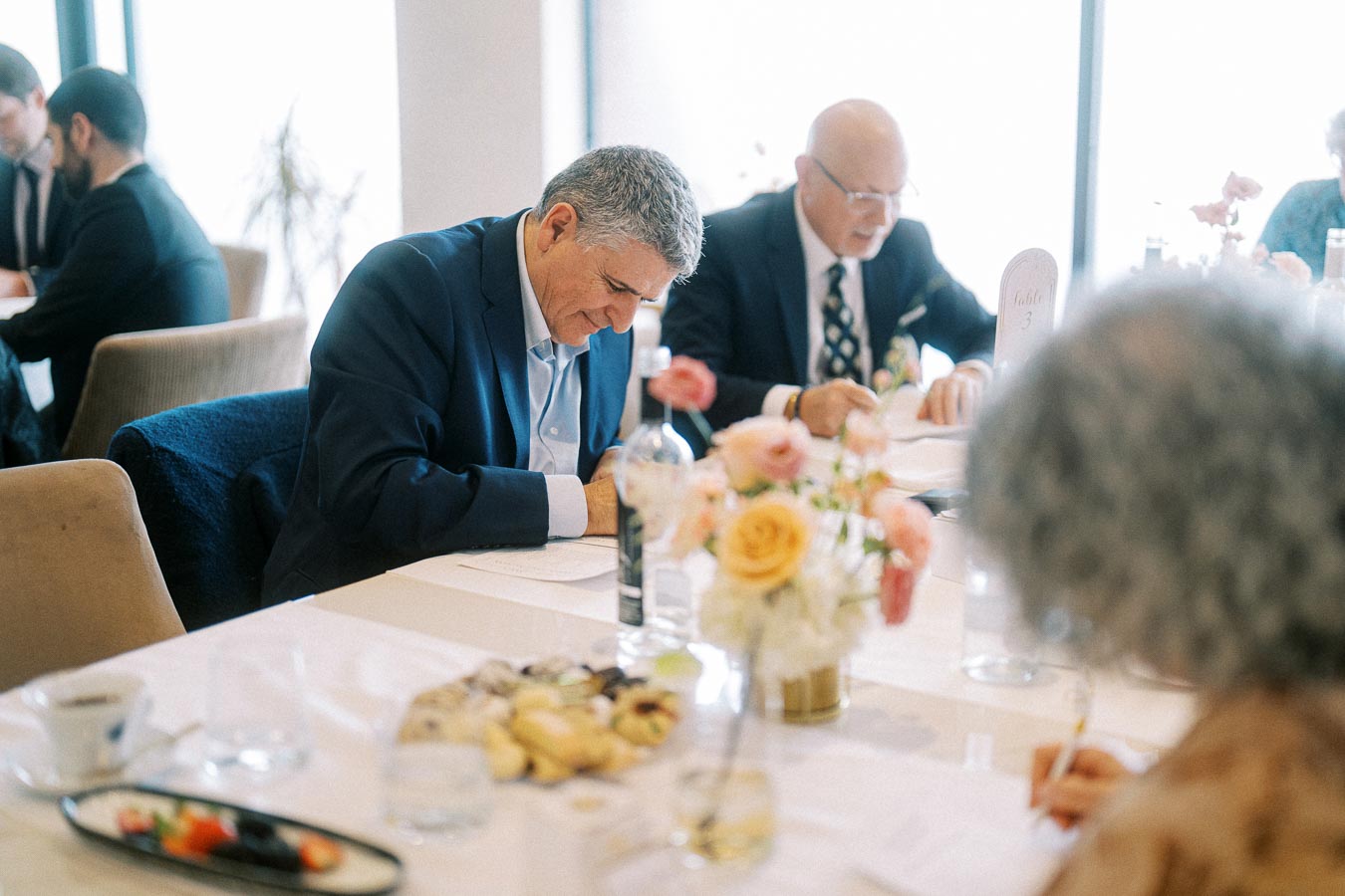 Elderly men in suits writing at a conference table with flower centerpiece and refreshments.