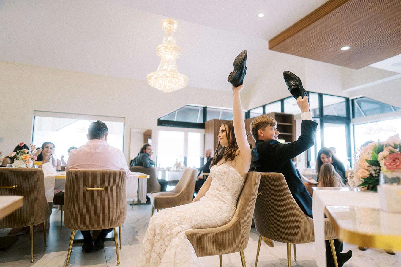 Bride and groom participating in a wedding shoe game, each holding up a shoe, inside a decorated reception venue with guests