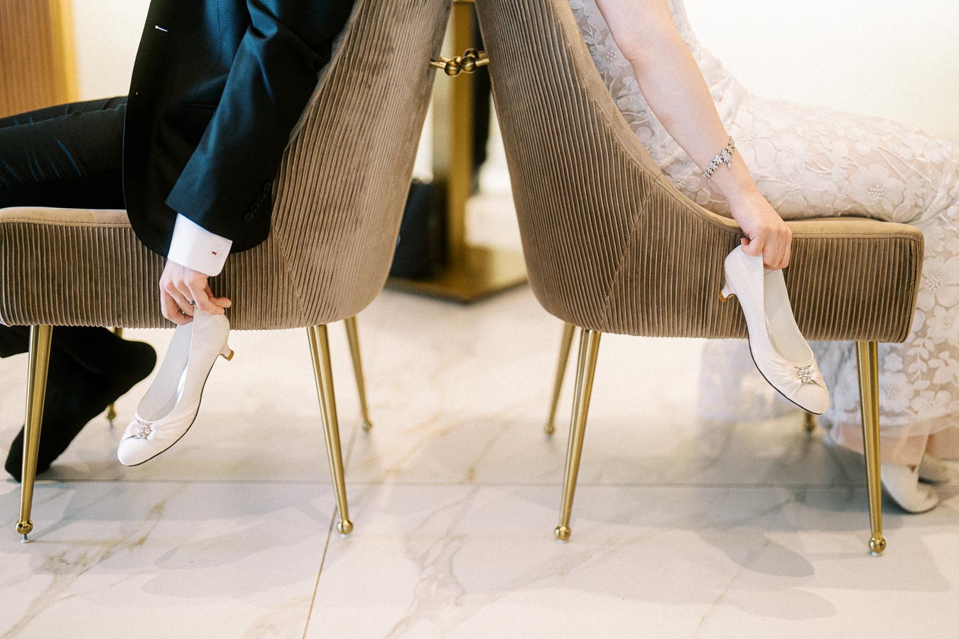 Bride and groom seated back-to-back, holding their shoes, during a playful wedding game in an elegant setting.