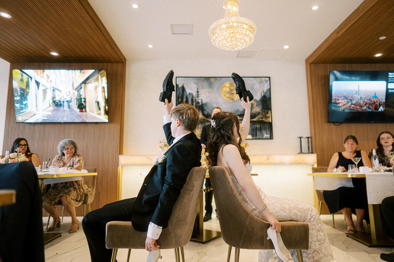 Bride and groom playing the shoe game at their wedding reception, sitting back-to-back and holding up shoes, with guests
