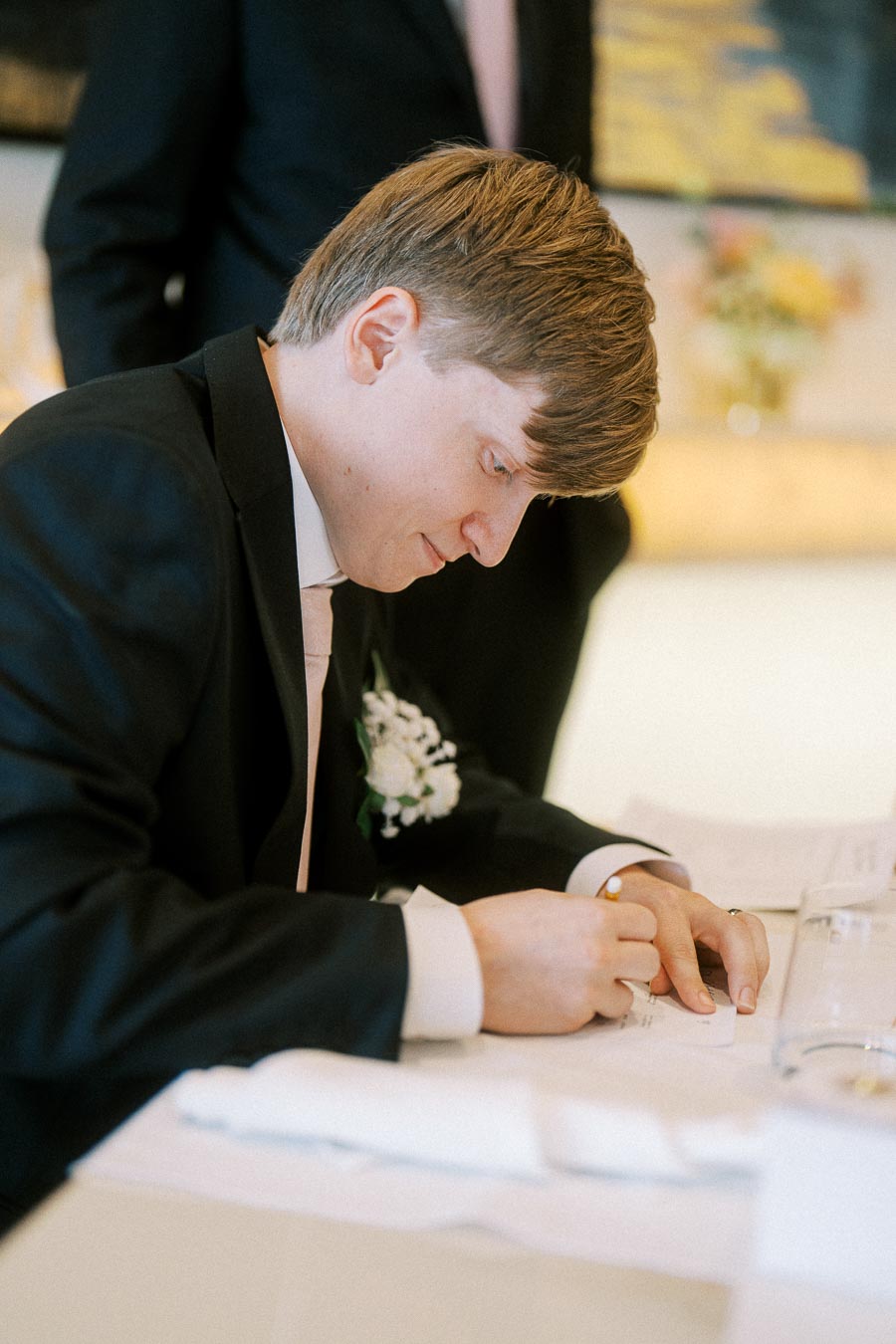A person in a formal suit signing a document, possibly during a ceremonial or official event, with a boutonnière on the