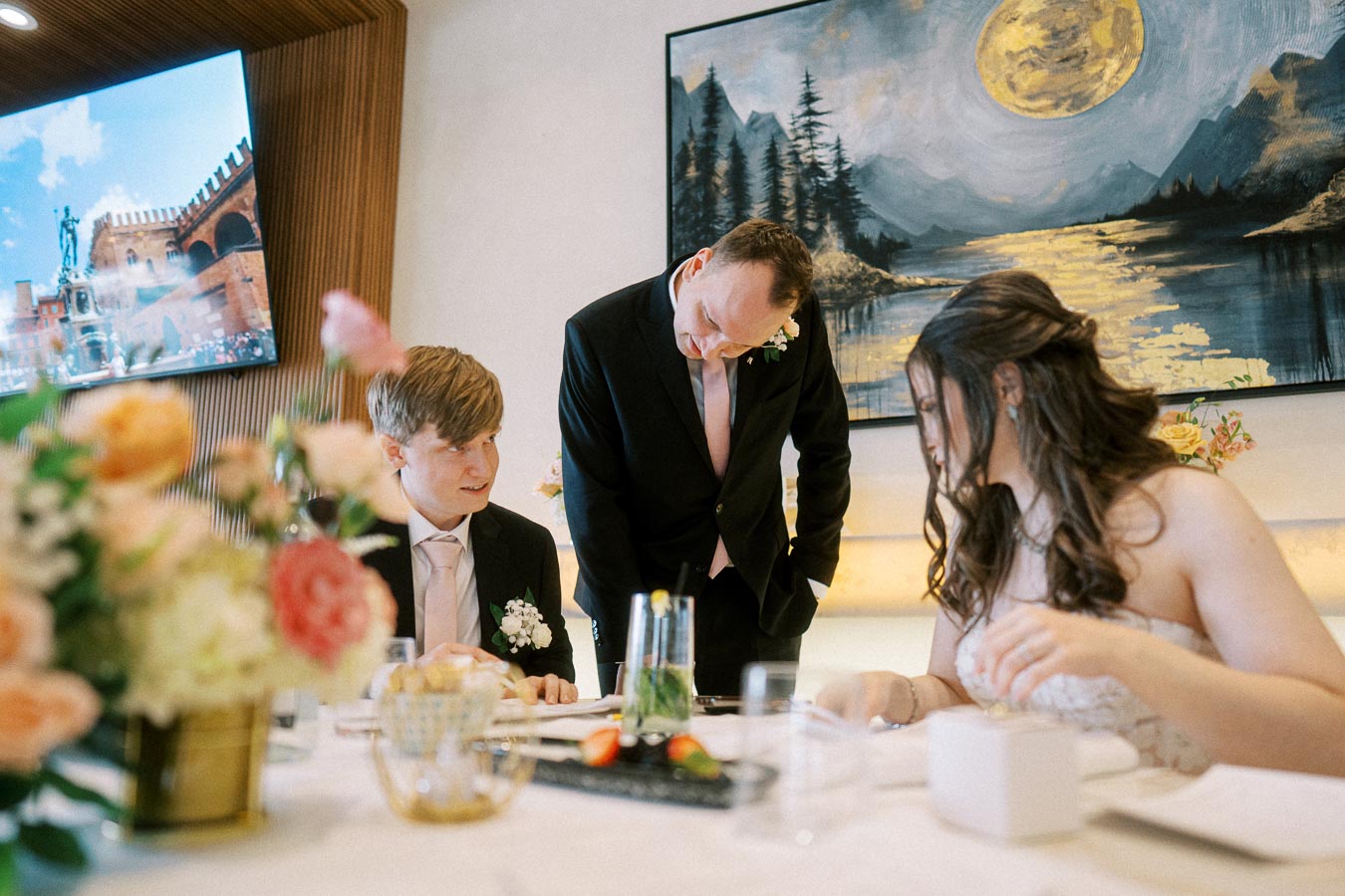 A bride and groom seated at a wedding reception table as a man in a suit leans over, engaged in conversation; artistic