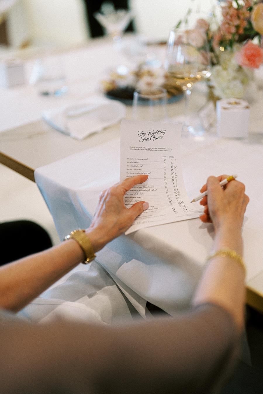 Person holding a pencil and engaging in the Wedding Shoe Game, surrounded by elegantly arranged table settings with flowers