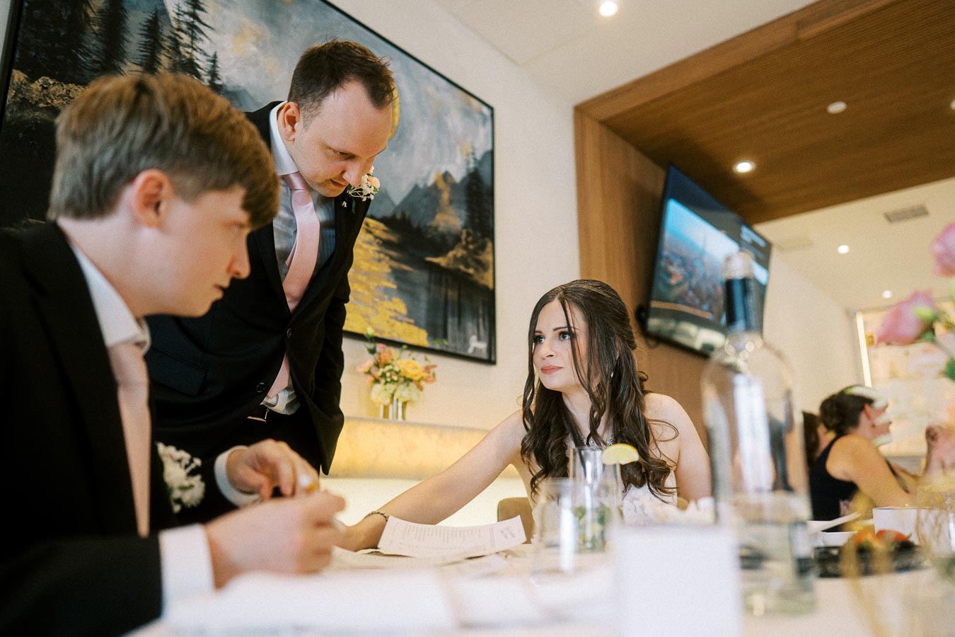 Group of people in formal attire at an indoor event, discussing documents at a table, with decorative flowers and a painting