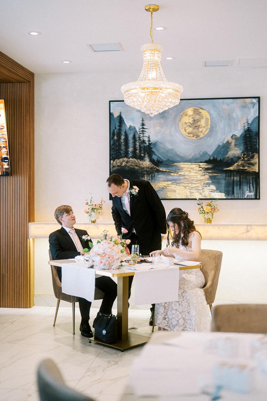 Elegant wedding reception scene with a couple seated at a dining table, adorned with floral arrangements. A painting of a