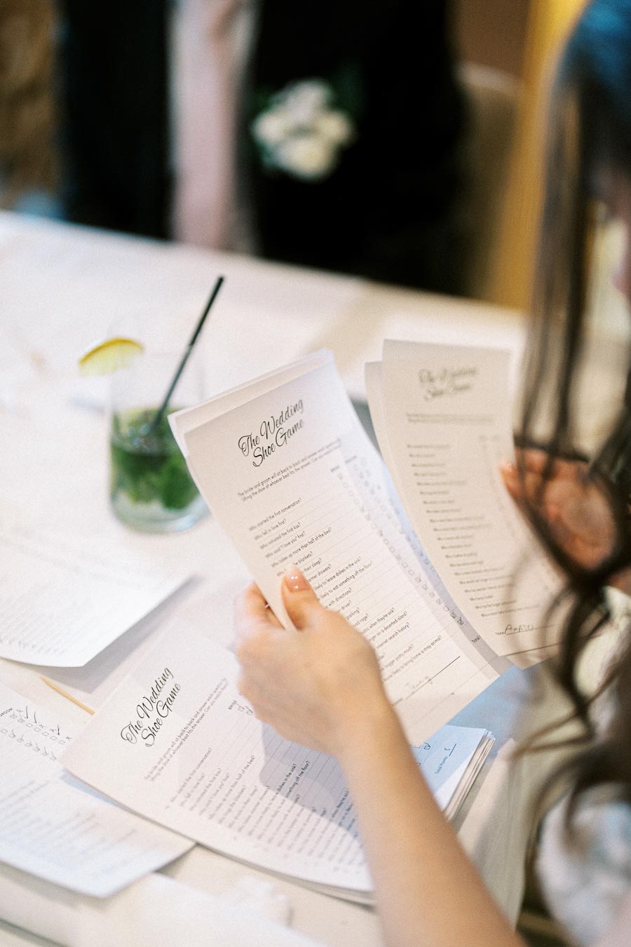 Person holding game cards for The Wedding Shoe Game at a formal event, with a cocktail on the table nearby.