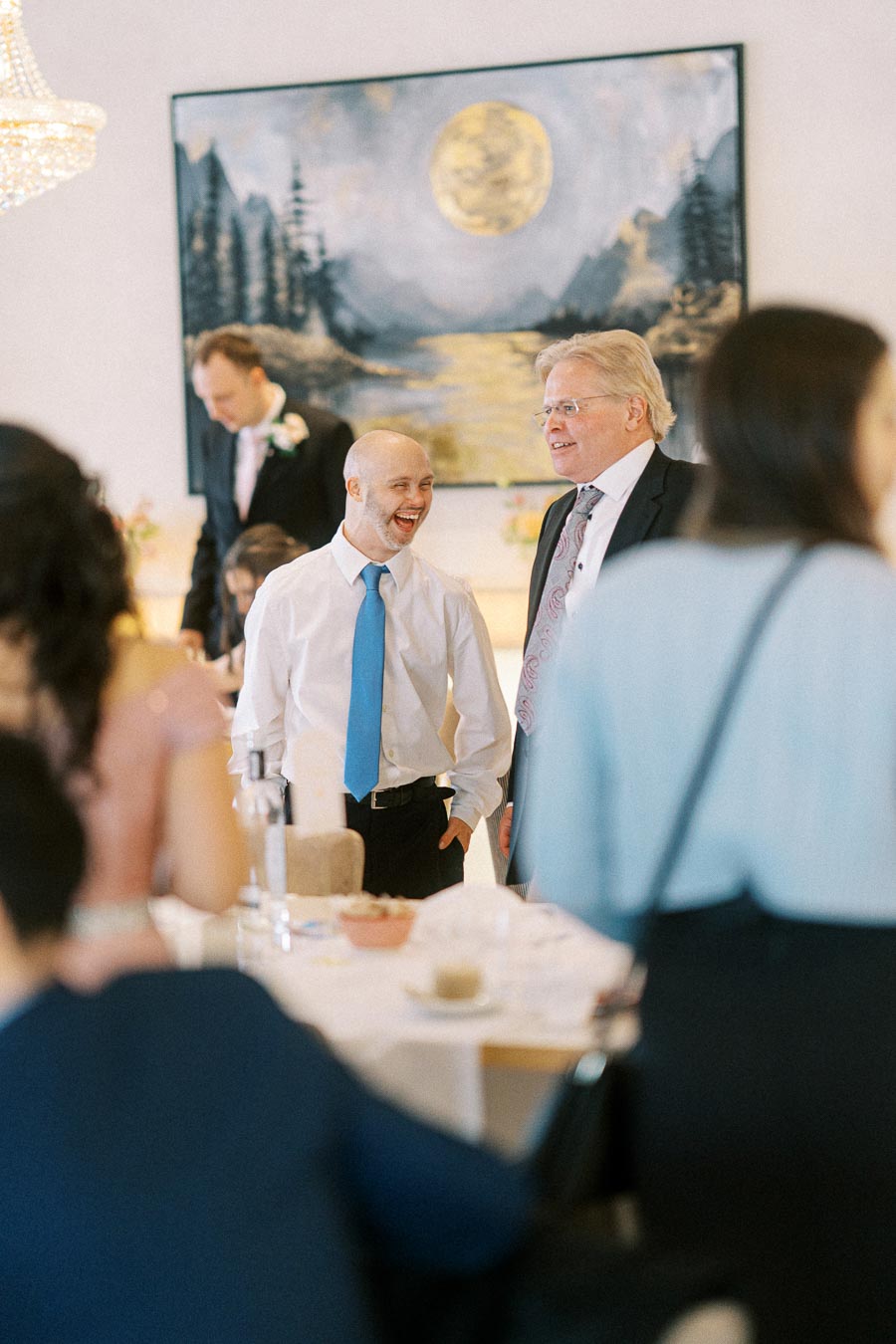 Group of people socializing at an indoor event, with a painting of a moonlit landscape in the background.