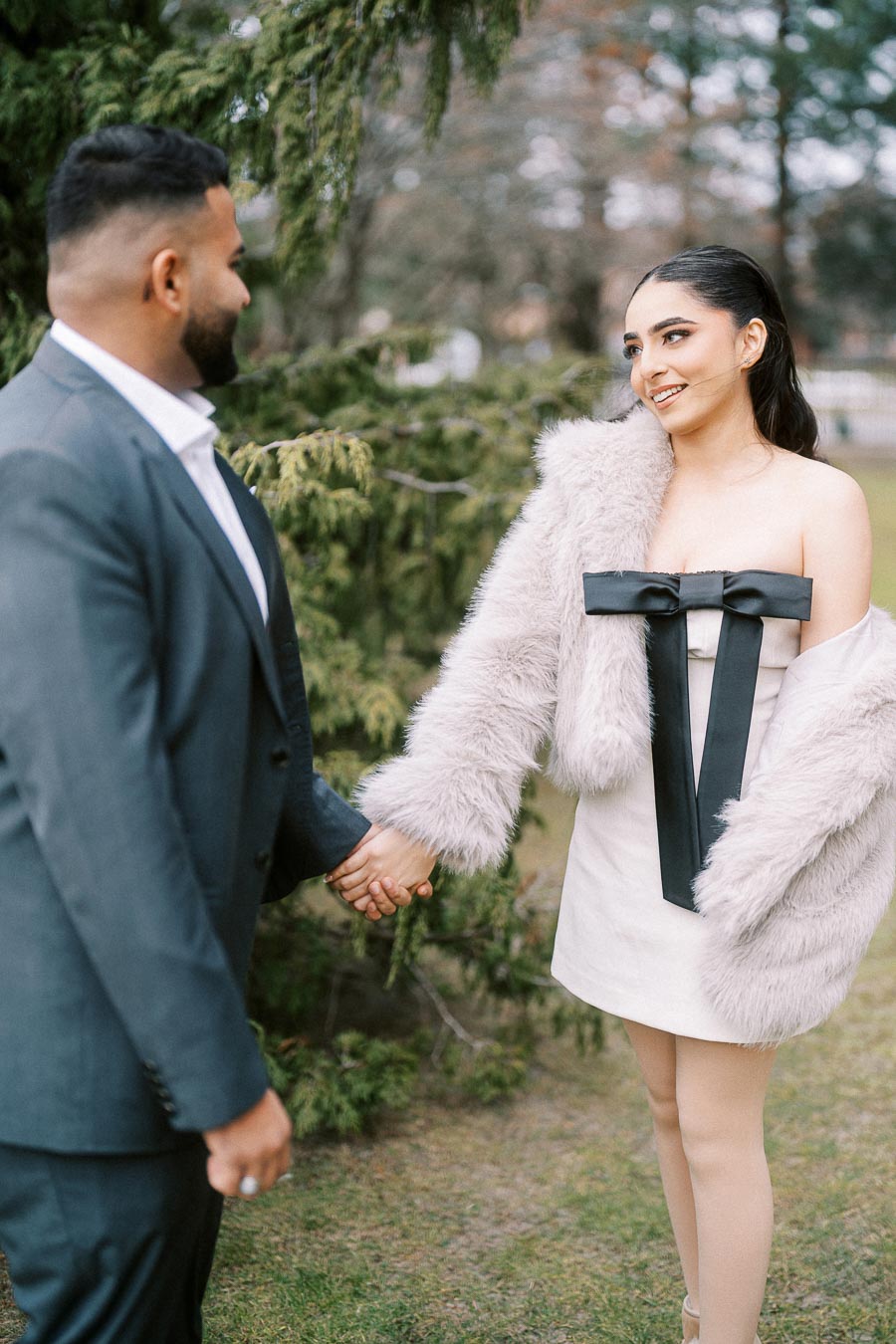 A couple holding hands outdoors, with the woman wearing a fur coat and decorative dress while the man is in a suit. Background features lush green trees, suggesting a serene and romantic setting.