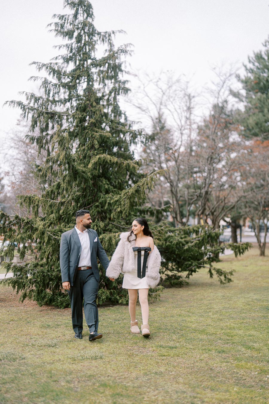 A couple walking hand in hand through a park, wearing stylish outfits; the man in a suit and the woman in a fur coat with a dress, with evergreen trees and a cloudy sky in the background.