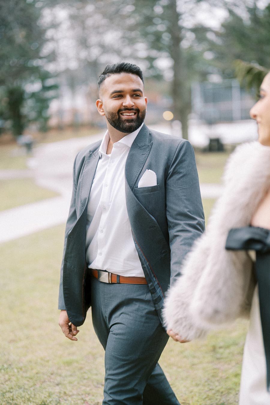 A man in a gray suit smiling while walking outside in a park setting, holding hands with another person. Trees and outdoor paths are visible in the background.