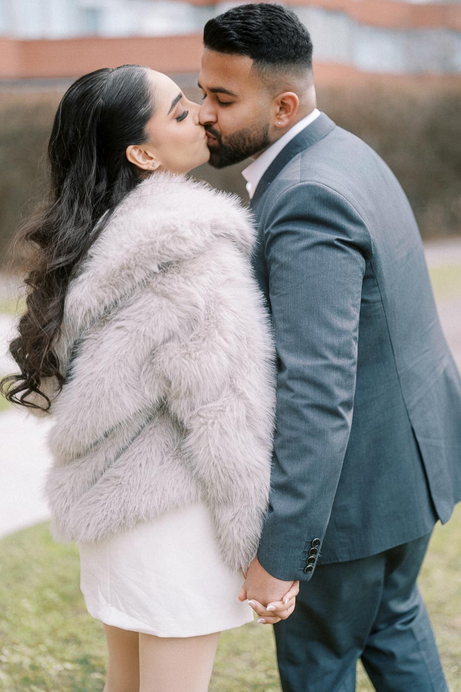 A couple sharing a romantic kiss in outdoor setting. The woman is wearing a stylish fur coat, while the man is dressed in a sharp gray suit. They are holding hands, capturing an intimate moment.