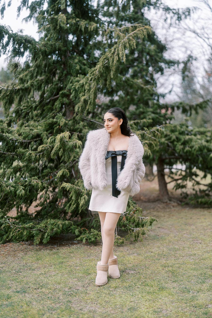 Young woman in a chic dress and faux fur coat standing in front of a pine tree in a park setting, exuding fashion-forward style with neutral ankle boots.