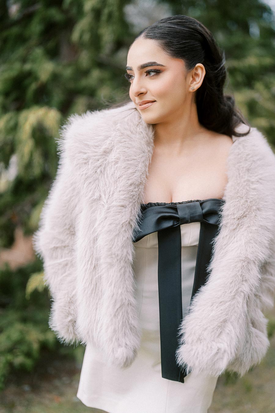 A woman wearing a fluffy light gray coat and elegant black and white dress with a bow detail, confidently posing outdoors with greenery in the background.