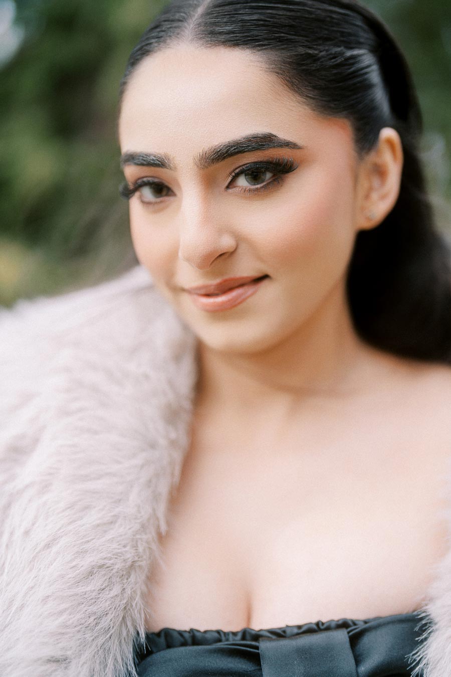 A woman with smooth dark hair is wearing elegant makeup, a soft fur shawl, and a stylish black dress, posing outdoors with a gentle smile.