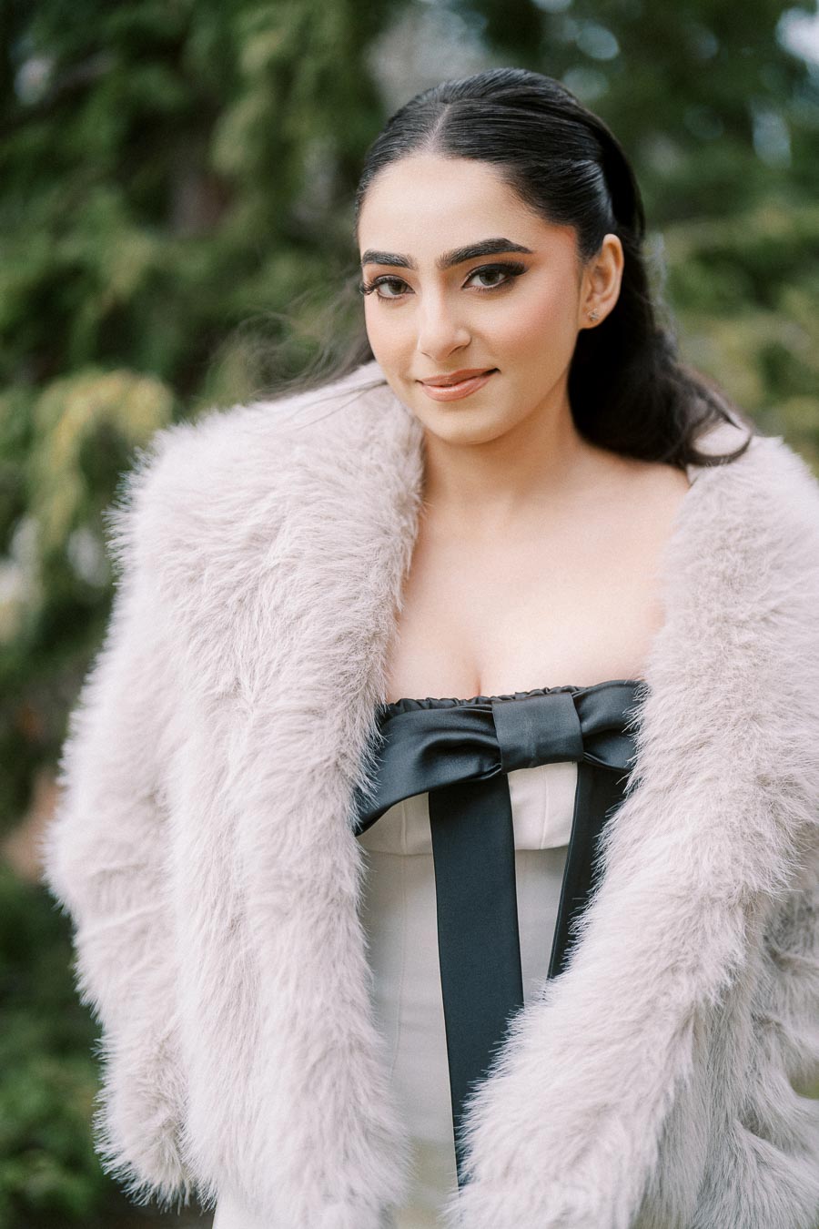 Young woman in a fluffy white coat and black bow dress posing outdoors with a natural background.