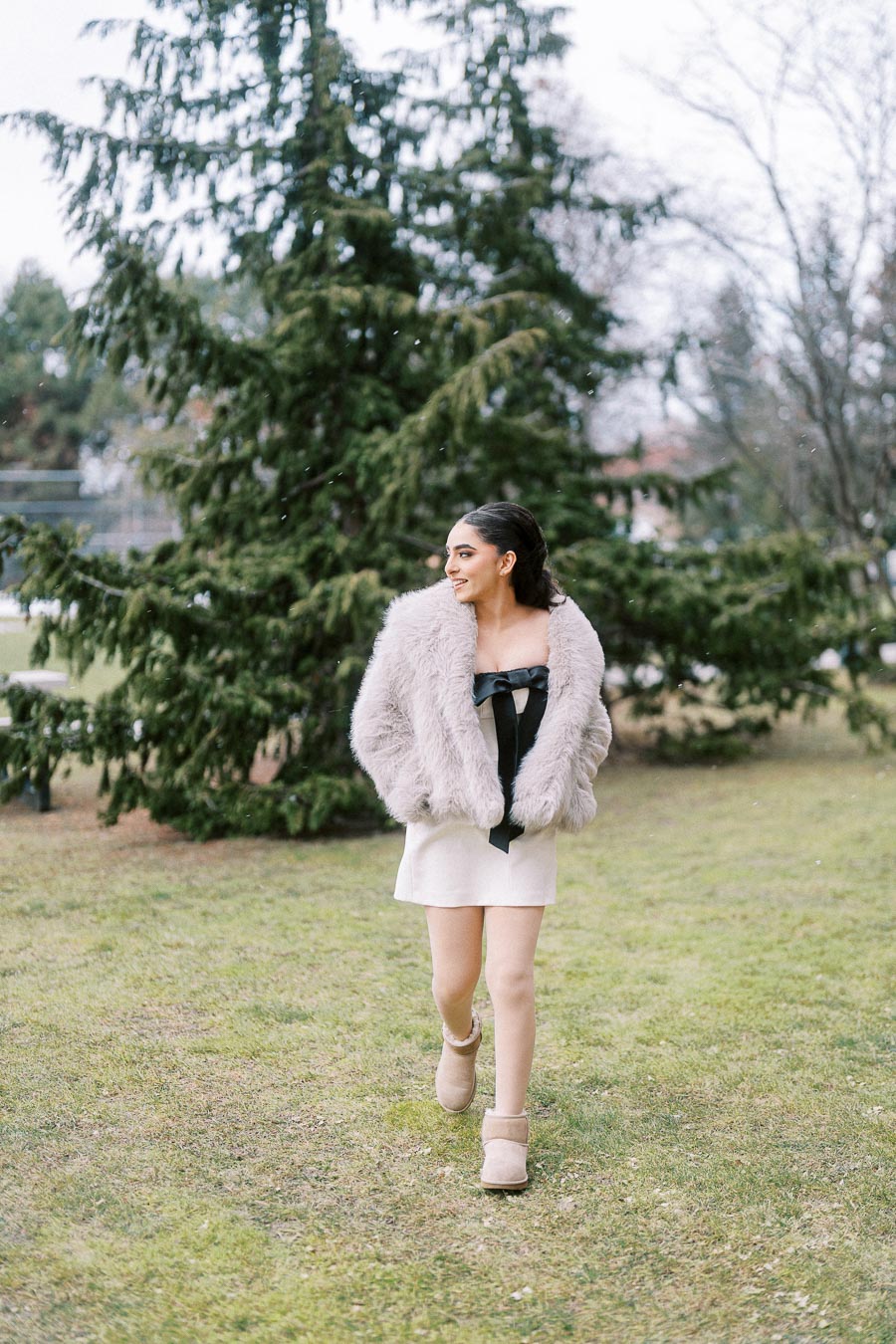 A stylish woman wearing a fur coat over a black dress walks on a grassy field with snowflakes falling, surrounded by evergreen trees in a winter setting.