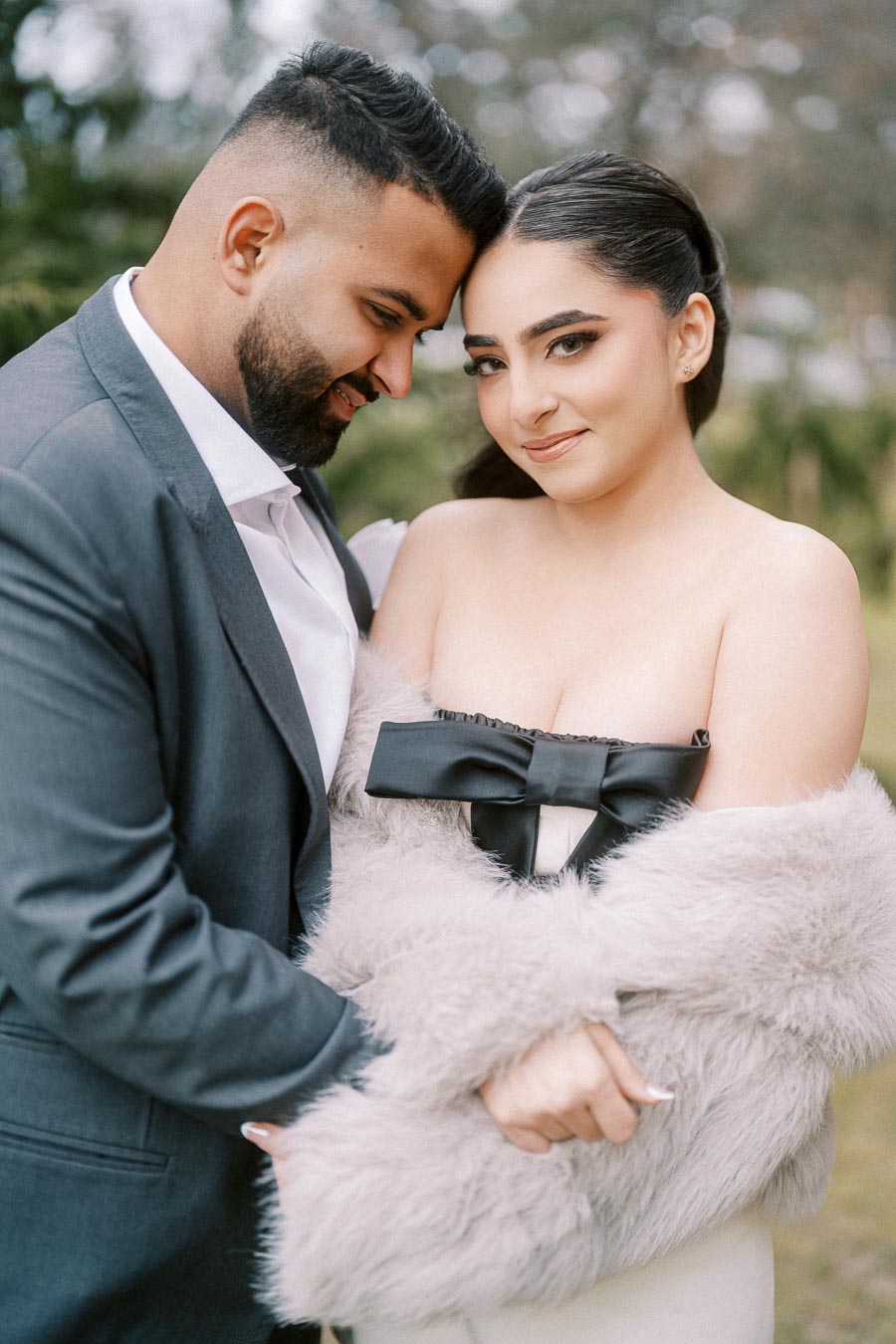 A smiling couple embraces outdoors, the woman wearing a fur shawl and elegant dress with a black bow, and the man in a gray suit.
