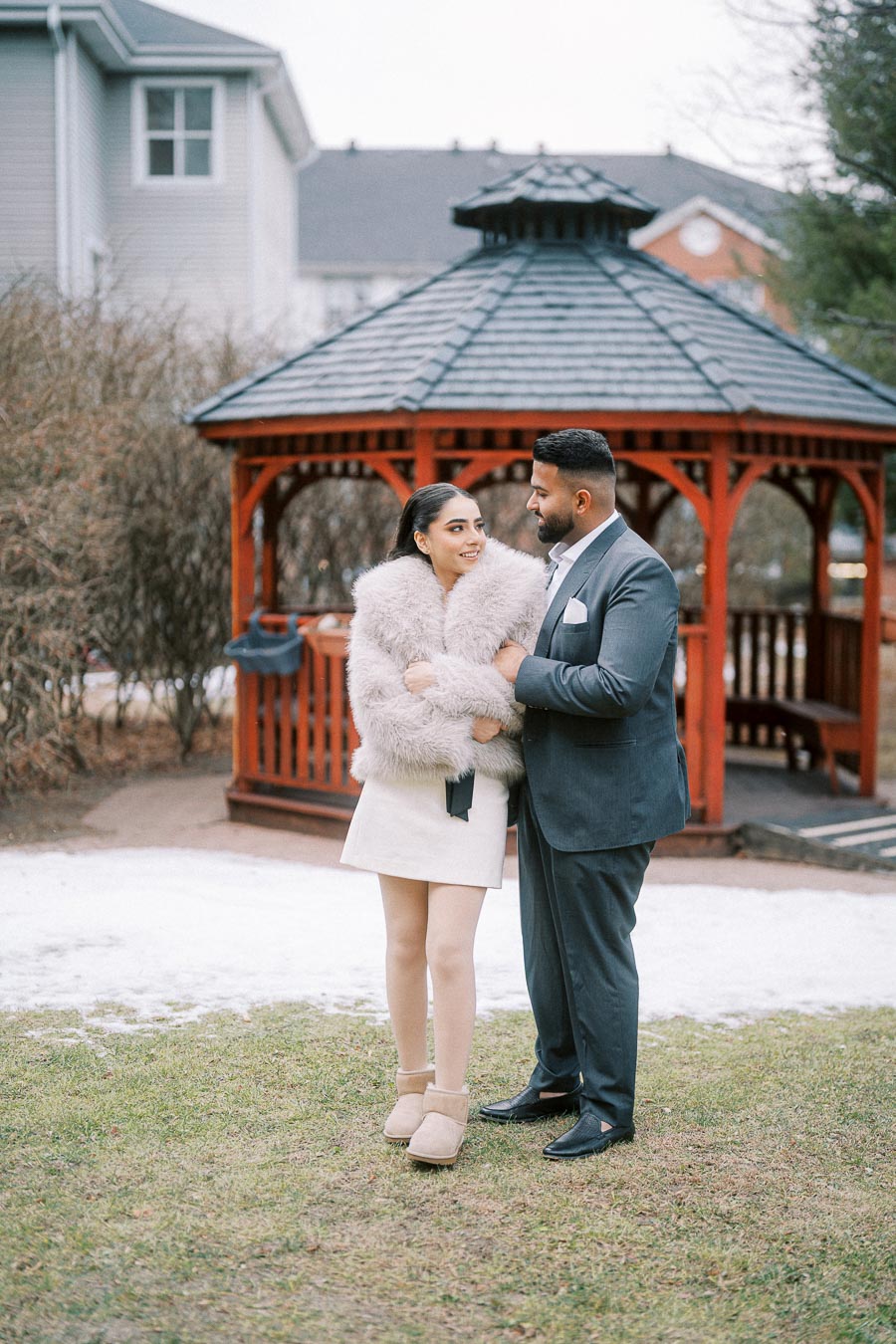 A couple dressed warmly, standing and smiling in front of a red wooden gazebo on a winter day, surrounded by snow and grass.