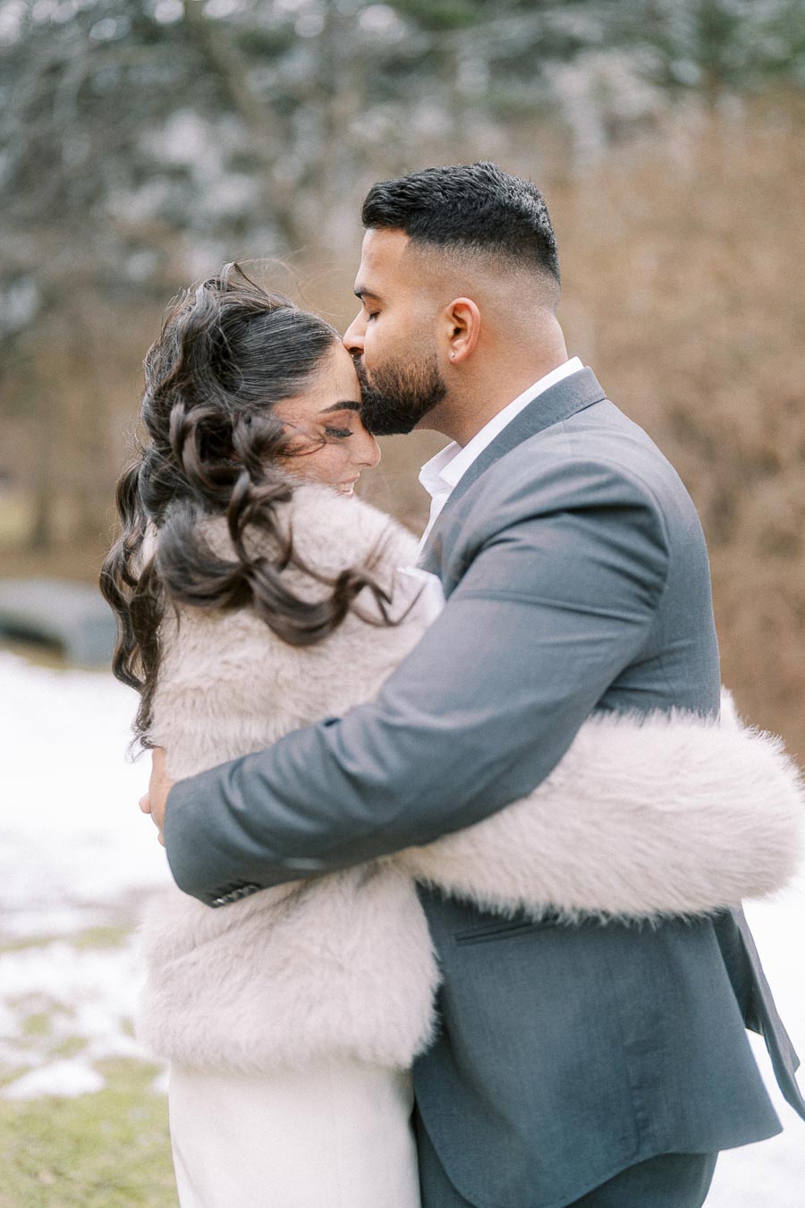 A couple embracing in a snowy outdoor setting, the man tenderly kissing the woman's forehead. The woman is wearing a fur coat, and the man is dressed in a suit, showcasing a romantic and intimate moment.