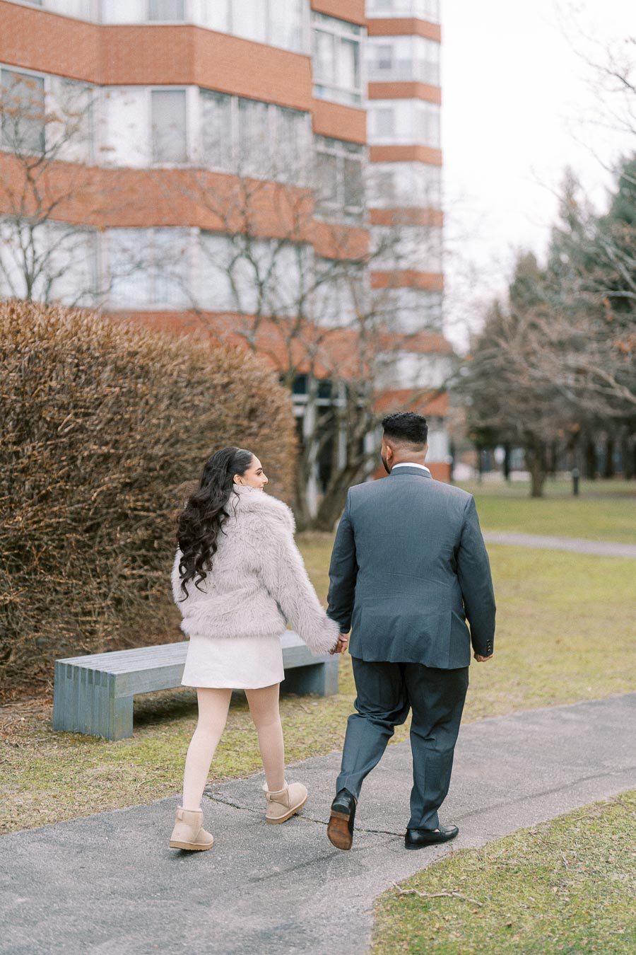 Young couple walking hand in hand on a path, surrounded by winter trees and an urban building backdrop.