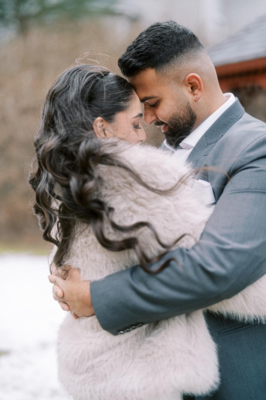 A couple embracing intimately outdoors in winter attire, with the man in a gray suit and the woman wrapped in a beige fur coat, creating a romantic ambiance.