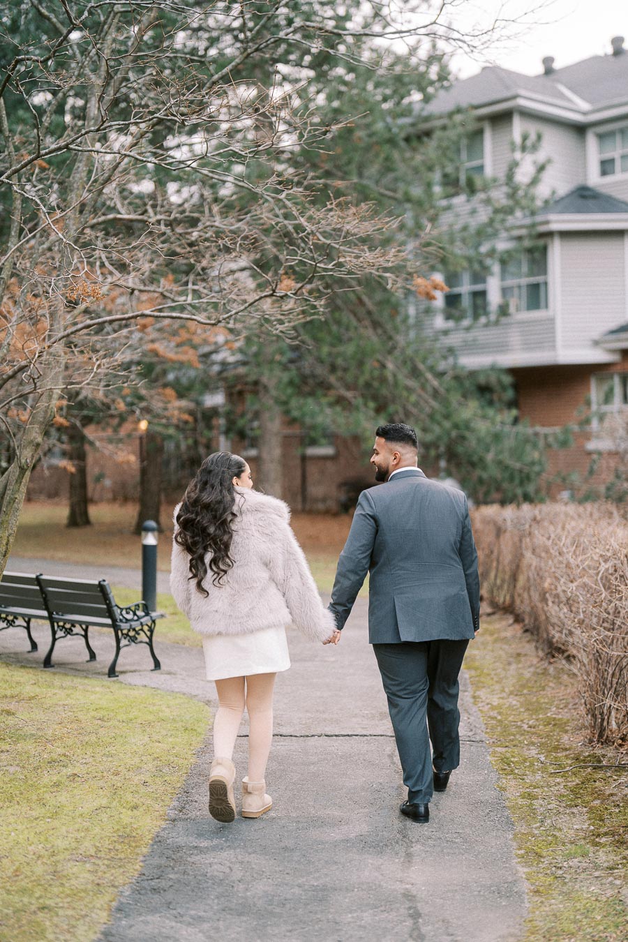 A couple walking hand in hand down a path, surrounded by trees and a residential building in the background, symbolizing love and togetherness on a peaceful day.