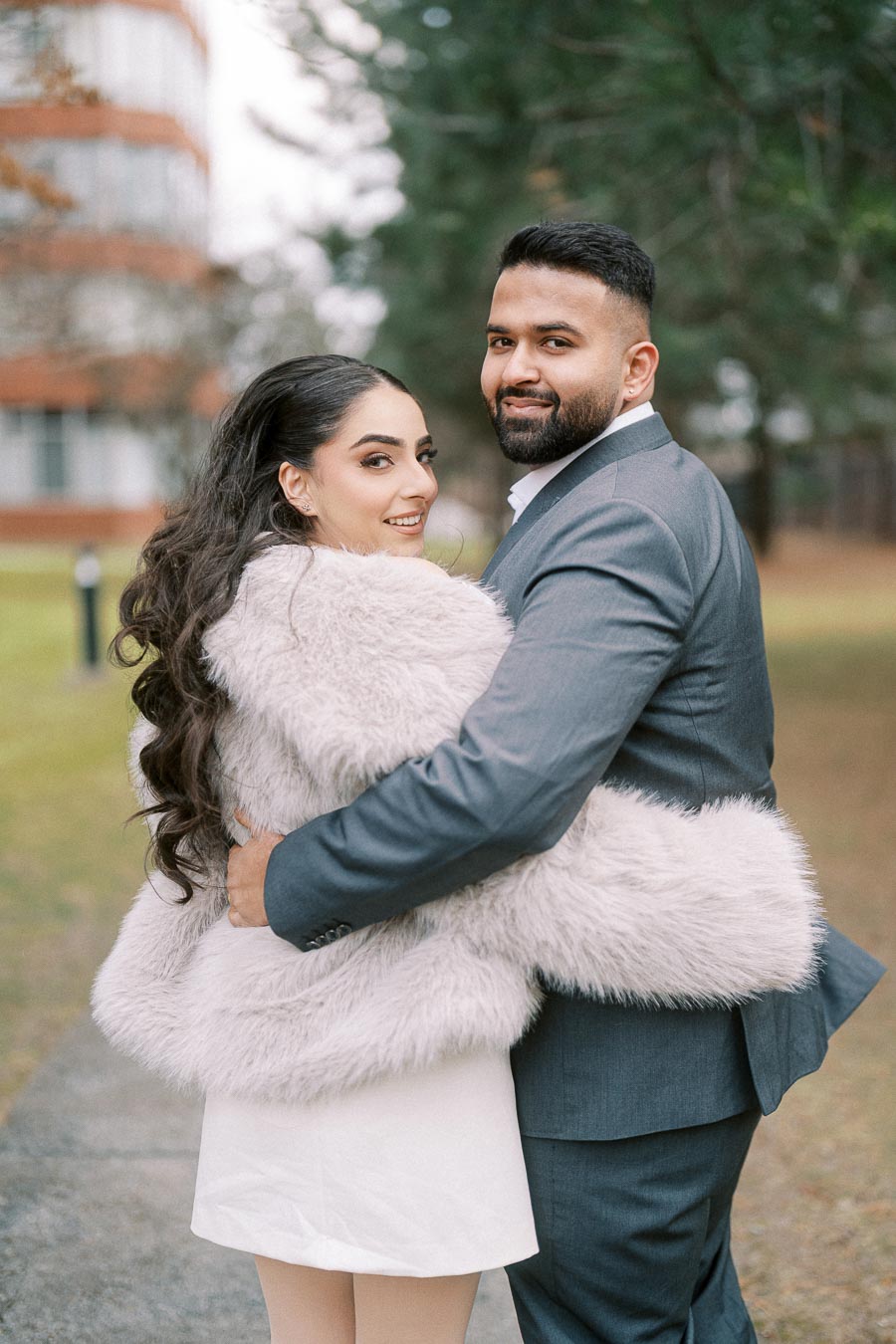 Couple embracing in an outdoor setting, with the woman wearing a fuzzy coat and the man in a gray suit, surrounded by greenery and a building in the background.