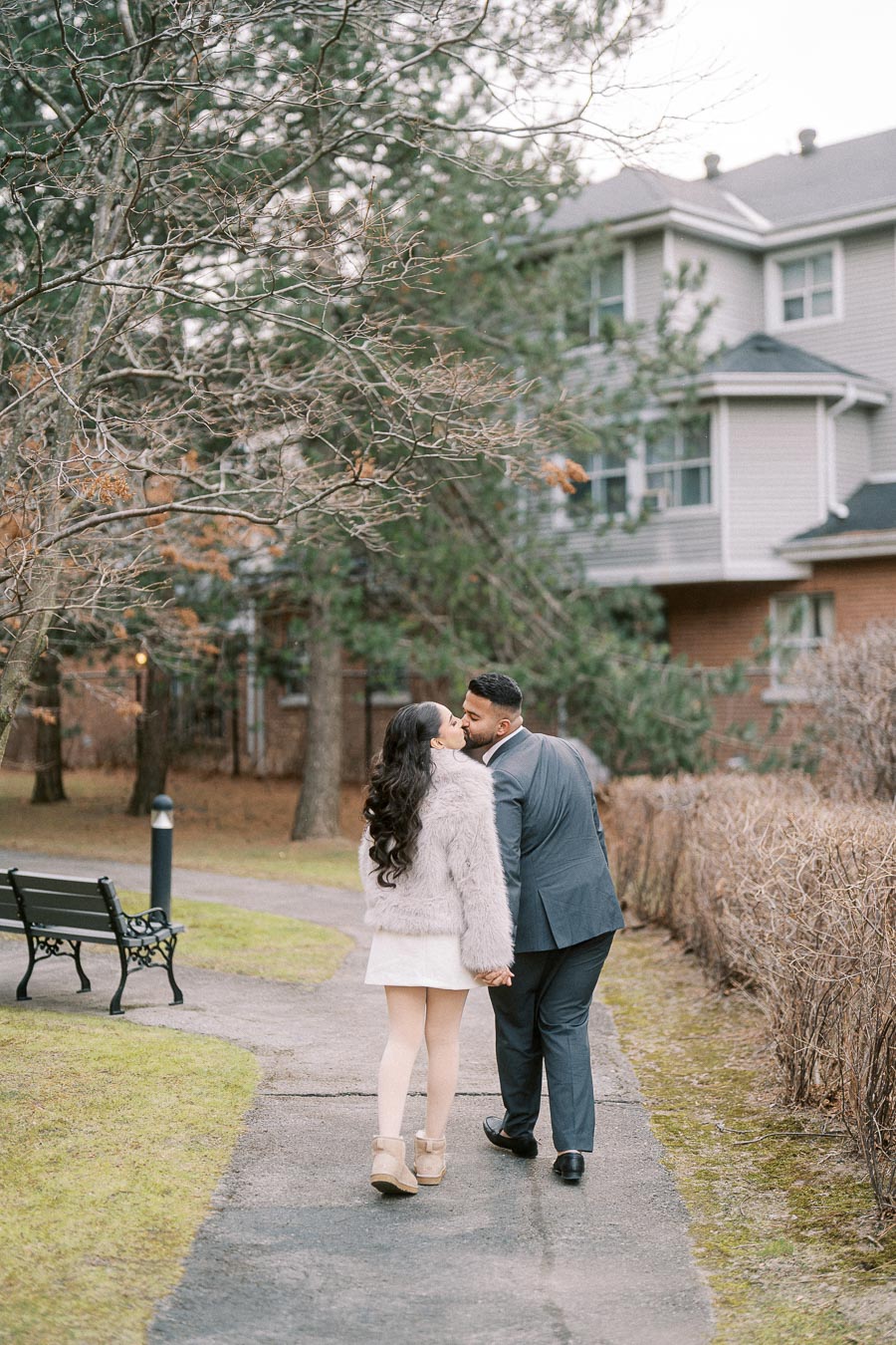 A couple walking hand in hand on a paved path through a park-like setting, surrounded by trees and shrubs, near a residential building. The woman is wearing a fur coat and boots, while the man is dressed in a suit.