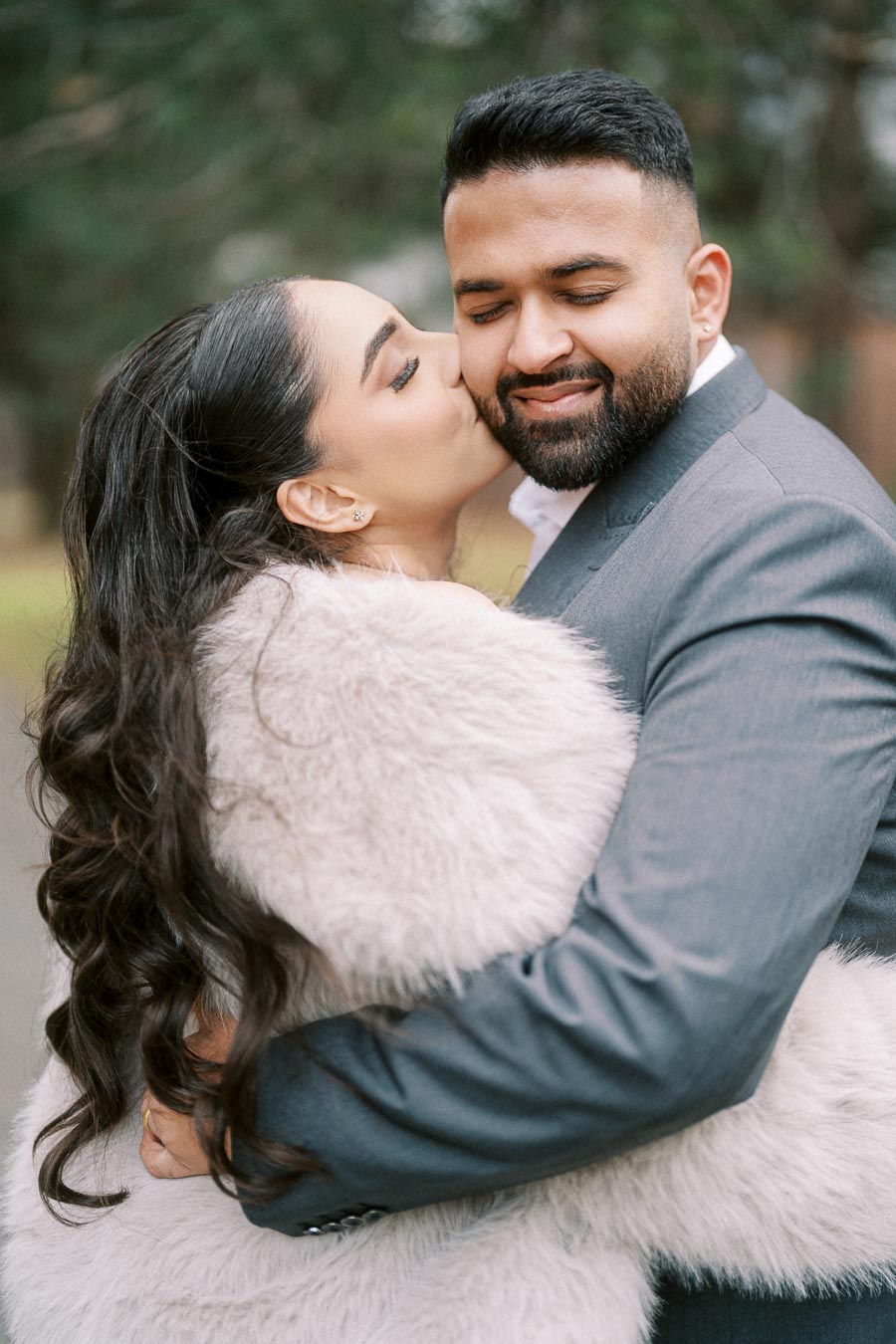 A couple embraces lovingly in an outdoor setting, with the woman kissing the man's cheek. She is wearing a fur coat, and he is dressed in a gray suit, both appearing joyful and content.