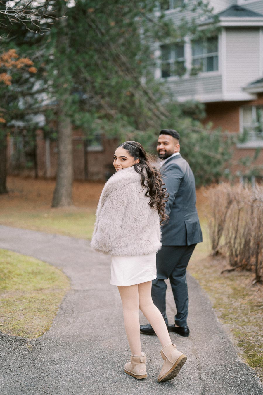 A couple walking along a tree-lined path, with the woman wearing a fluffy gray coat and beige boots, and the man dressed in a sharp gray suit, both looking back towards the camera, smiling, with a residential building in the background.