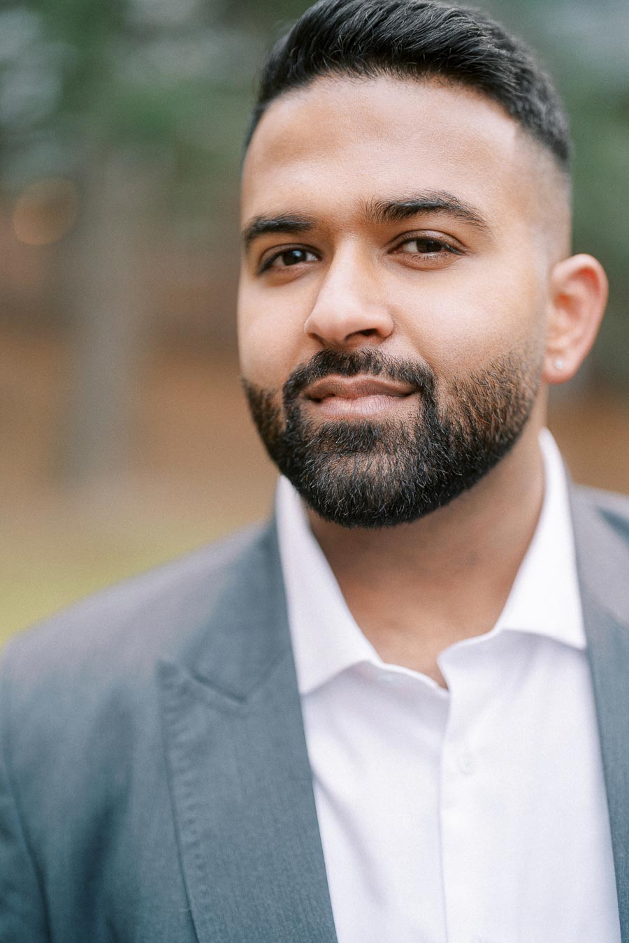 Portrait of a man with a beard wearing a gray suit jacket and white shirt, outdoors with a blurred natural background.