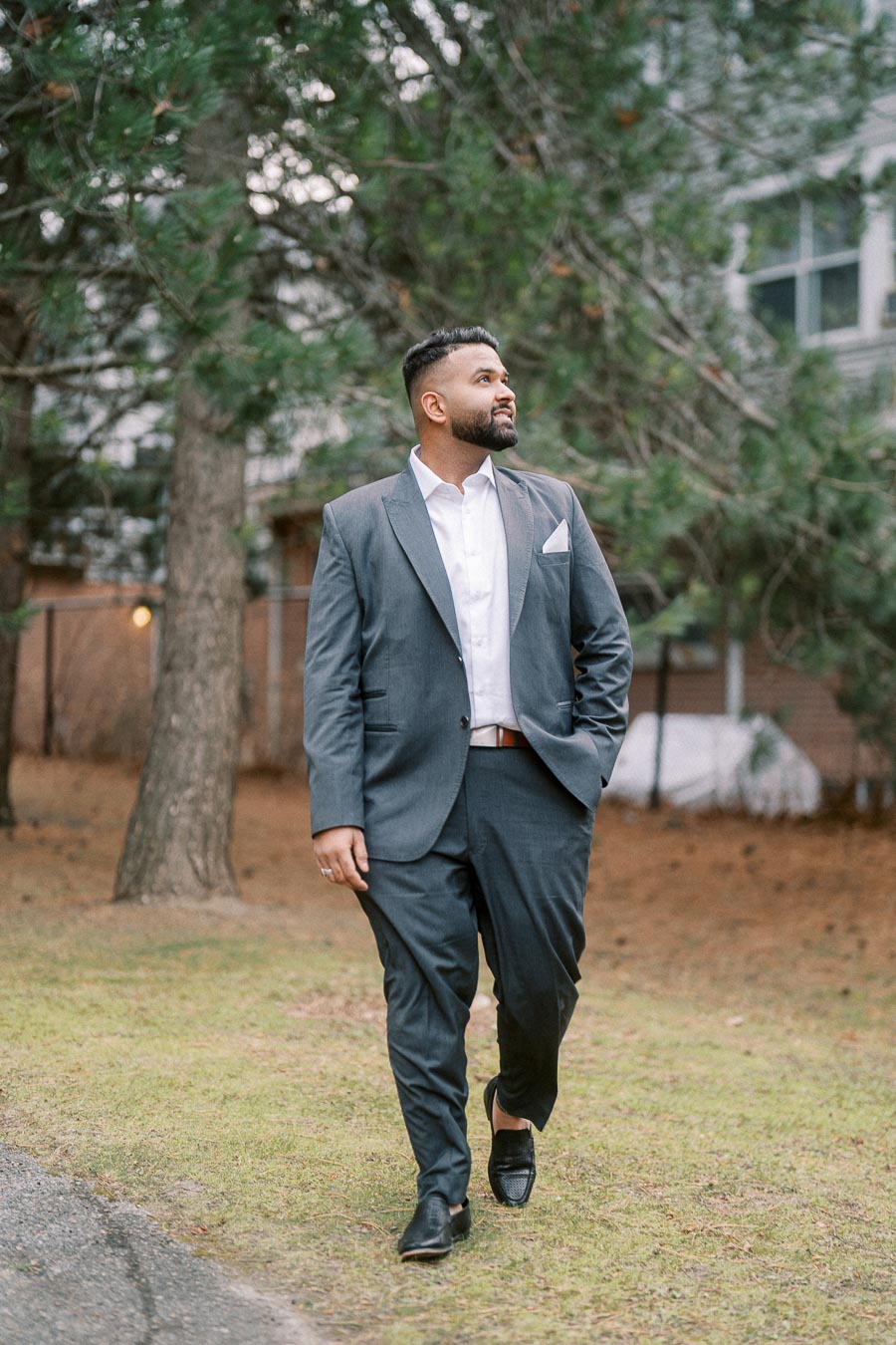 A man in a stylish gray suit walks confidently on a grassy path, with trees and a building in the background.