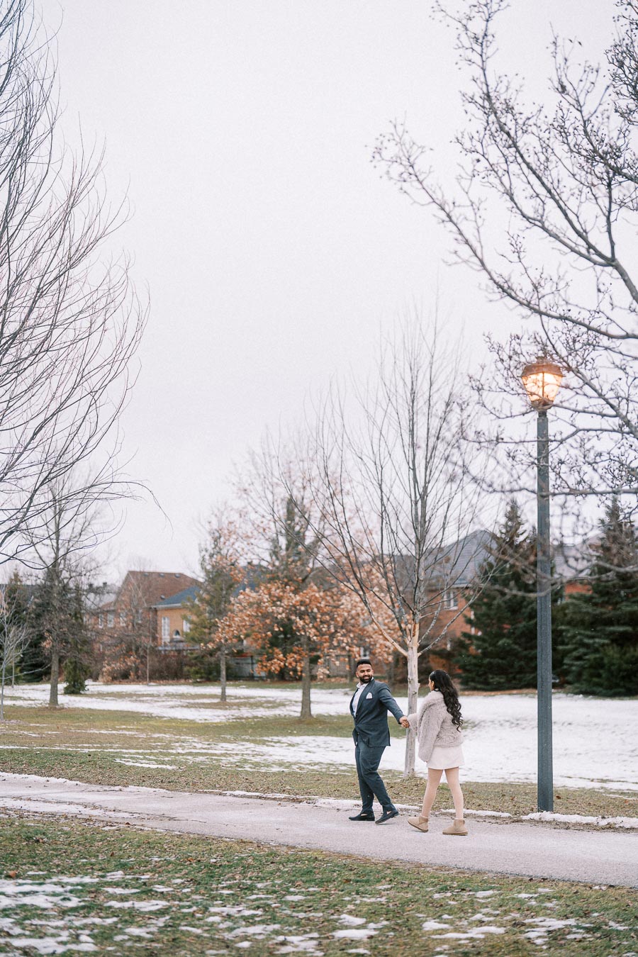 A couple holding hands and walking on a snowy park path surrounded by barren trees and a lamppost, with houses in the background on a cloudy day.
