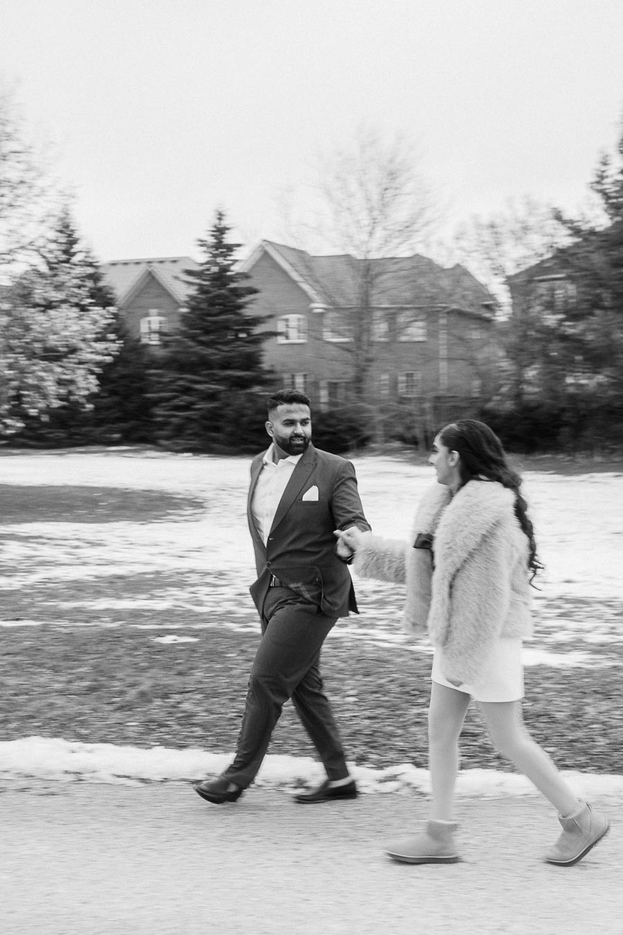 A couple walking hand in hand along a snowy pathway in a suburban neighborhood, with the man wearing a suit and the woman in a fur coat, showcasing a winter engagement photo in black and white.