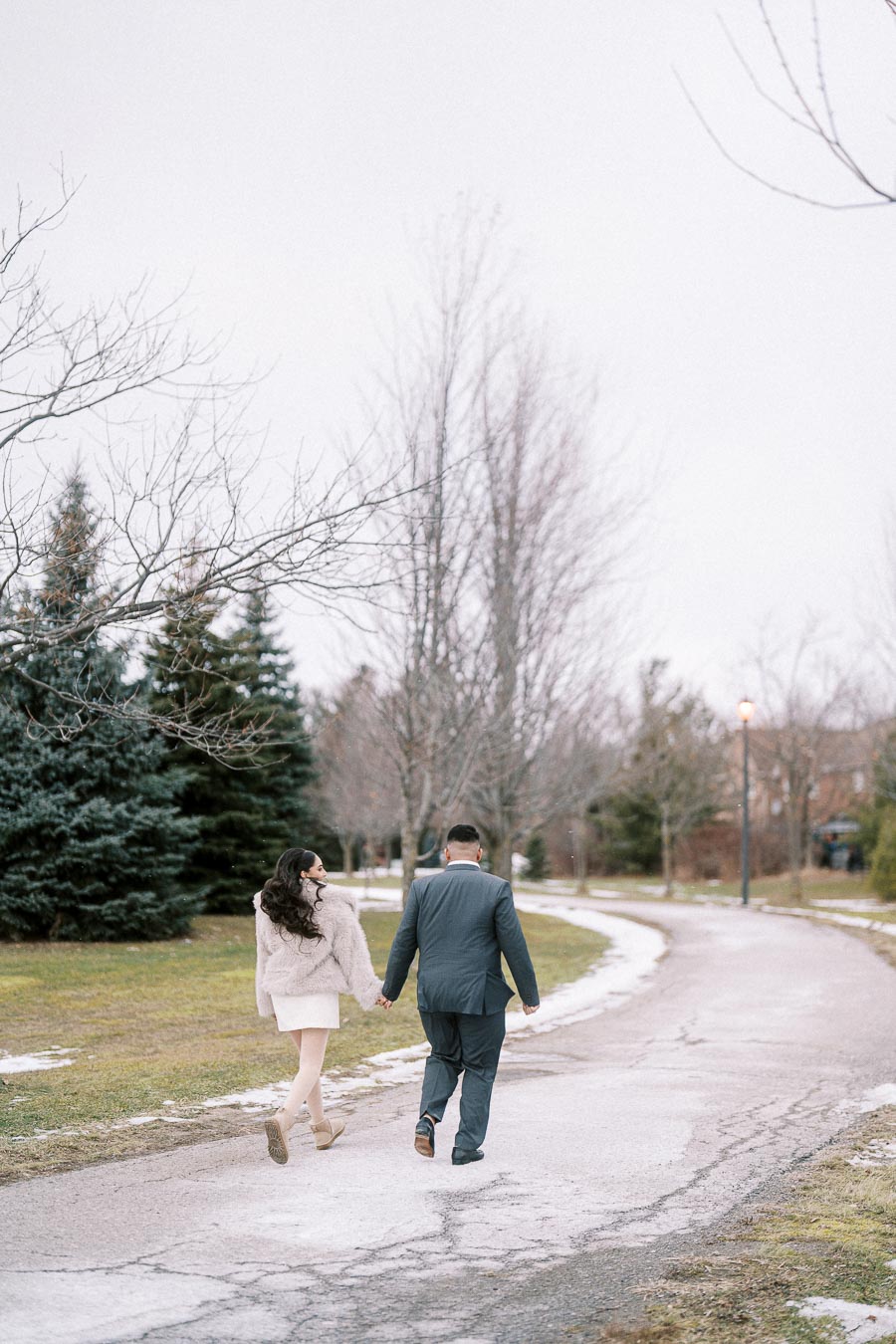 A couple holding hands walking down a snowy path in a park, surrounded by bare trees and evergreens, on a cold, overcast day.