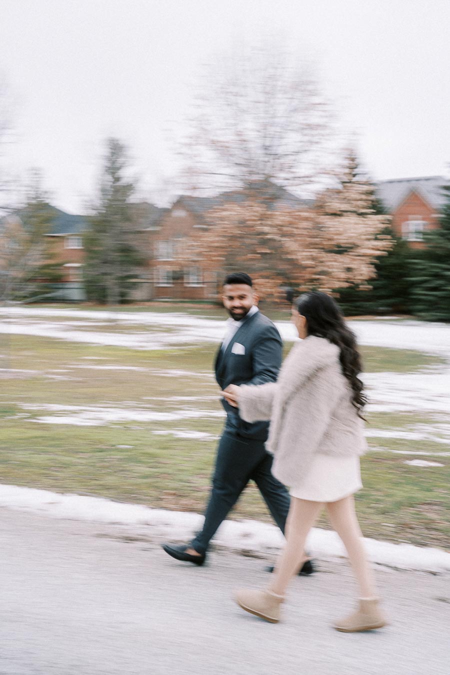 A couple walking hand in hand on a snowy day, with a suburban neighborhood and trees in the background. The man is wearing a suit and the woman is in winter attire, including a fur coat and boots.