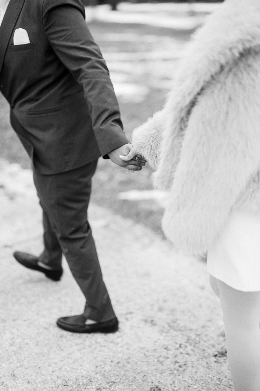 Black and white photo of a couple holding hands, with the man in a suit and the woman in a fur coat, walking outdoors on a snowy path.