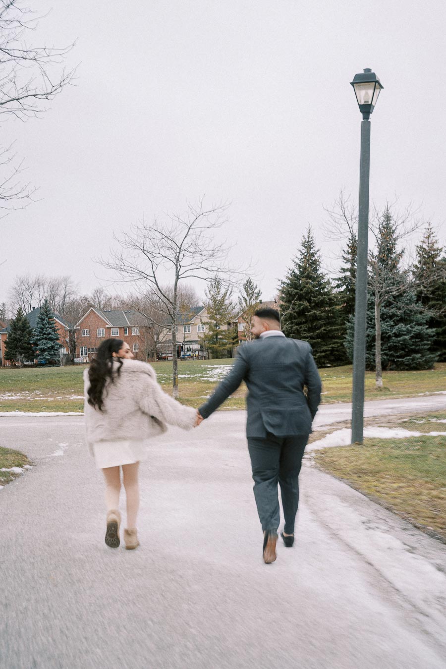 A couple walking hand in hand down a snowy path in a park, dressed warmly in winter clothing, with evergreen trees and houses in the background.
