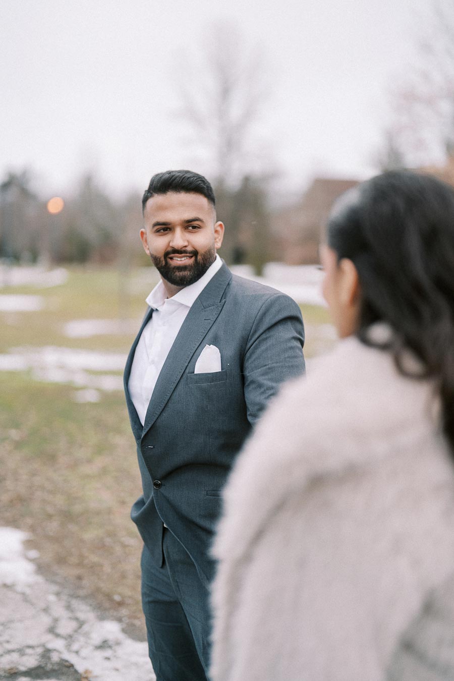 A man in a suit smiling while looking at someone outdoors in a park setting with sparse trees and patches of snow on the ground.