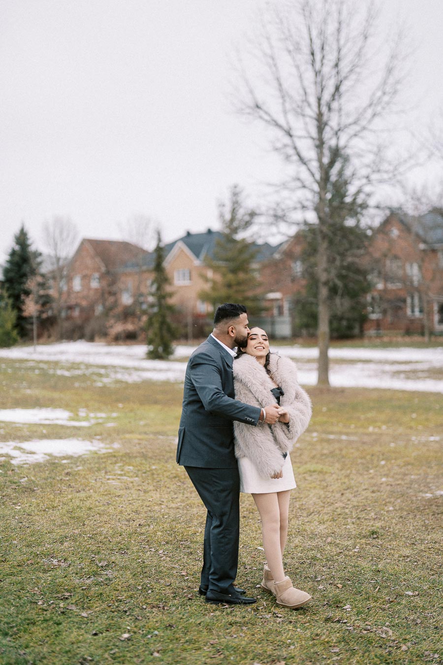A couple embraces warmly on a chilly day, standing on a grassy field with patches of snow, with houses and bare trees in the background.