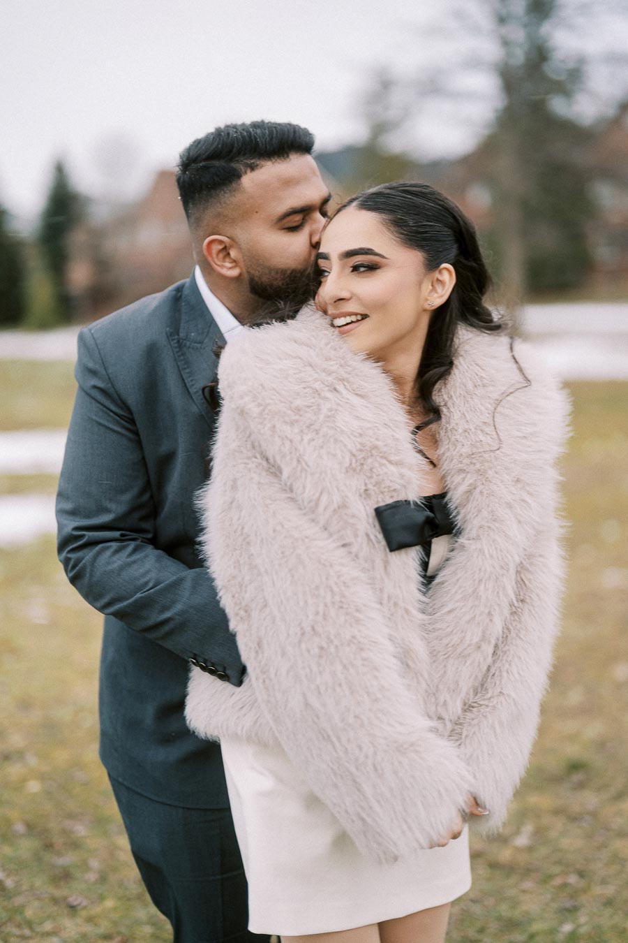 A couple in love poses outdoors, with a man in a suit kissing a woman wearing a fluffy coat, evoking romance and happiness.