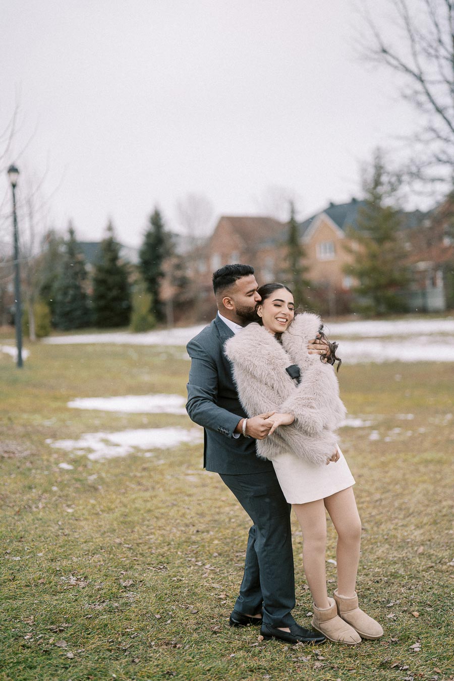 A couple enjoying a romantic moment outdoors in winter clothing, with the man embracing the woman from behind on a grassy field with trees and houses in the background.