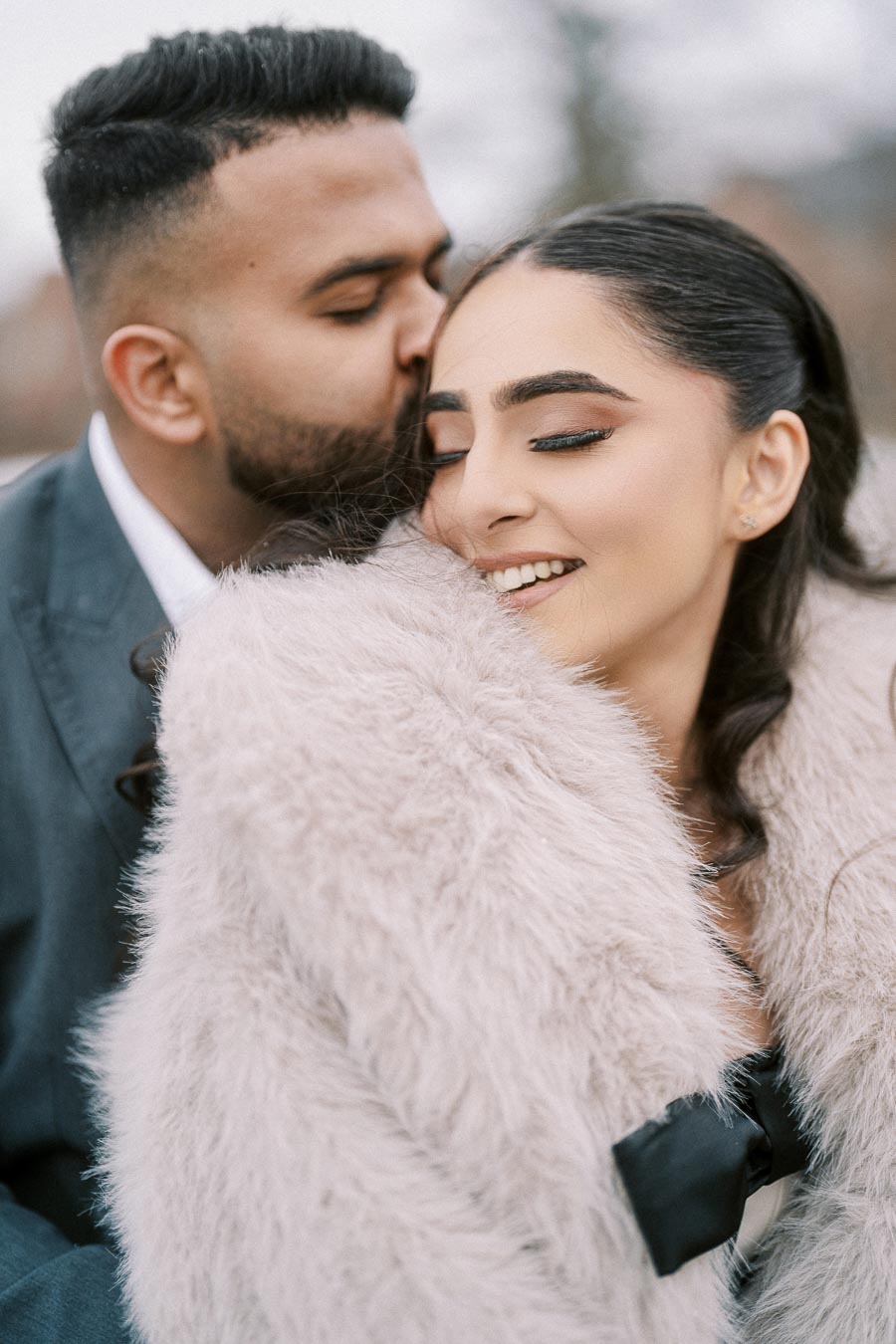 Romantic couple sharing an intimate moment with a kiss, woman in a fluffy coat smiling, close-up portrait outdoors.