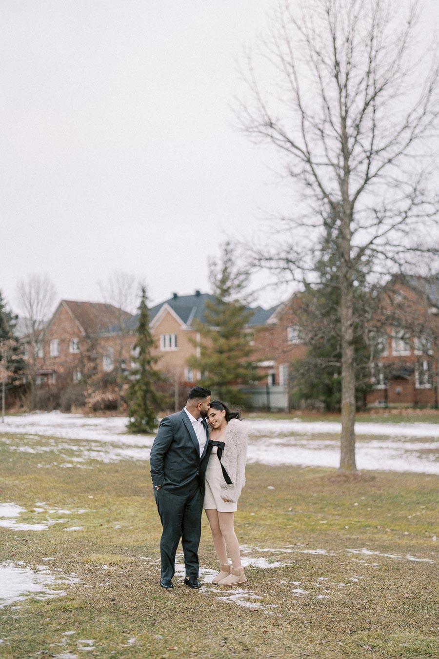 A couple standing in a snowy park, with the man in a suit and the woman in a dress and coat, leaning against each other affectionately with a backdrop of trees and houses.