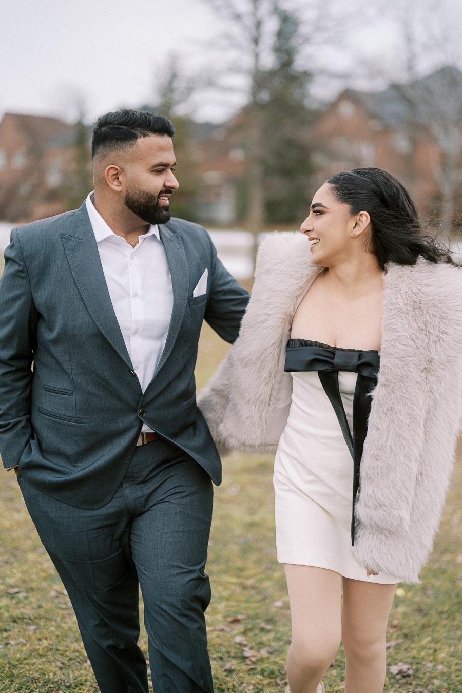 A couple walking outdoors in elegant attire; the man is in a gray suit, and the woman is wearing a white dress with a fur coat, both smiling and looking at each other.