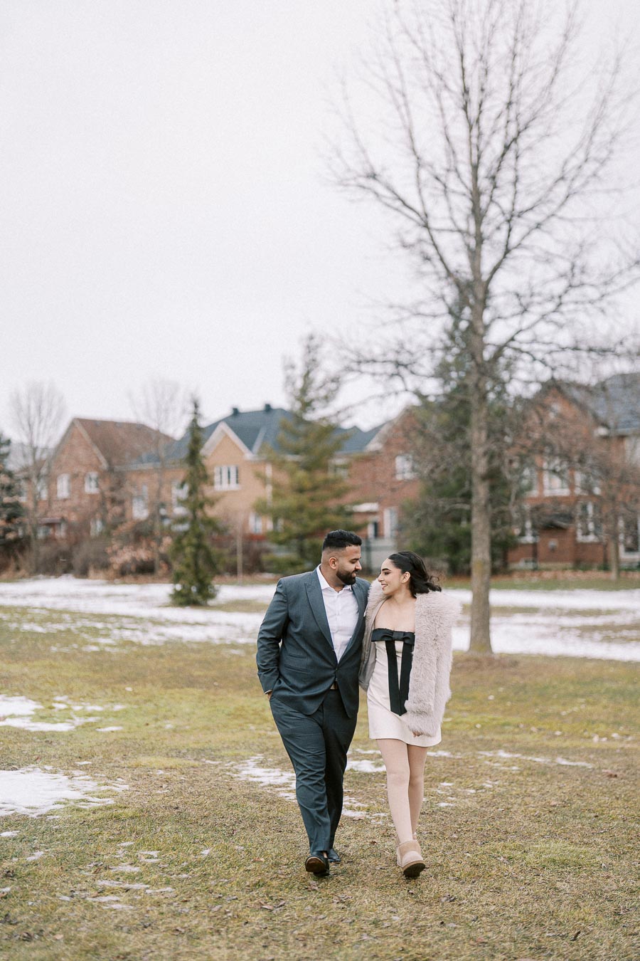 A couple walking arm in arm on a snowy day in a park with houses and trees in the background. The man is wearing a gray suit, and the woman is in a light dress and coat, showcasing a romantic winter setting.