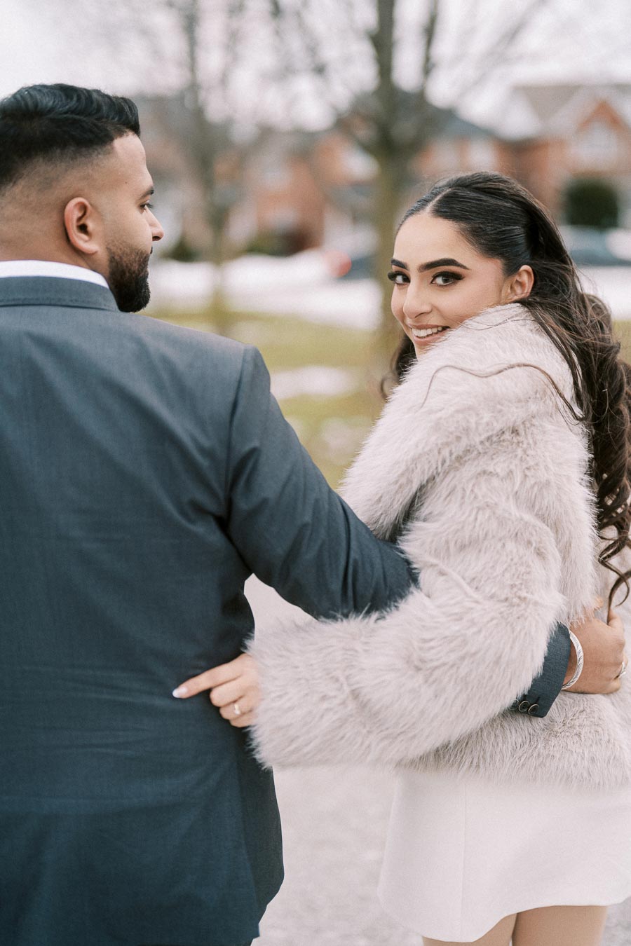 A couple walking arm in arm outdoors on a winter day; the woman is wearing a fluffy fur coat and smiling at the camera.