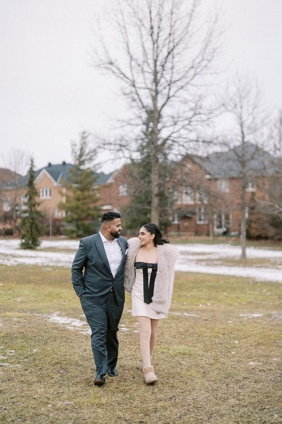 Couple walking together on a winter day in a park, dressed in elegant outfits, with residential houses and bare trees in the background.