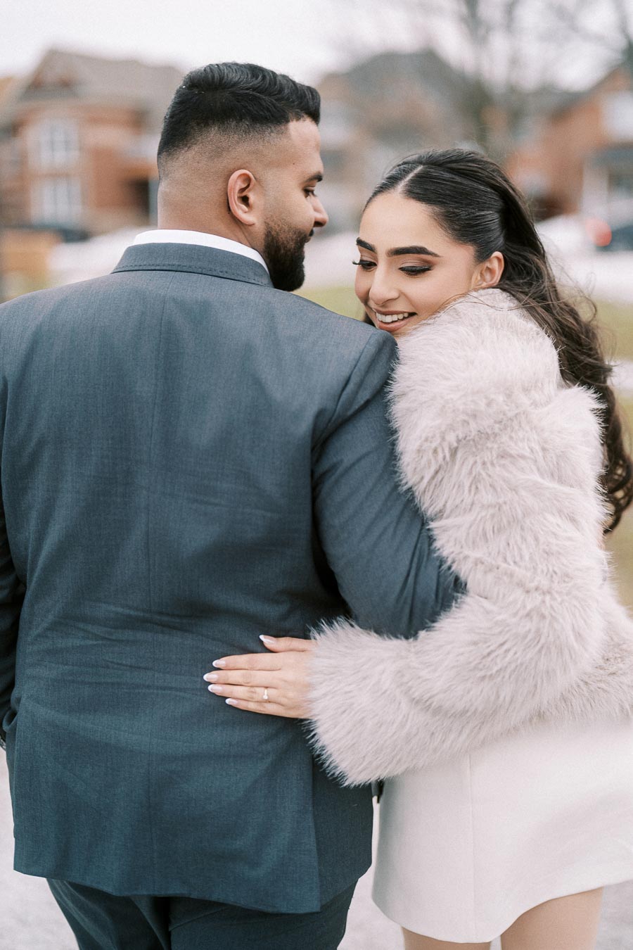 Couple embracing, woman in a white fur coat smiles affectionately at man in a suit, outdoors on a cloudy day.